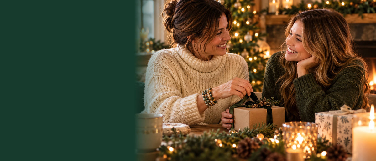 Two women sharing a wrapped gift beside a Christmas tree and a glowing fireplace.